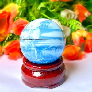 May include: A blue and white orb, resembling a miniature planet, rests on a small, round, red wooden stand. The orb's surface has a marbled appearance. The background features orange and yellow flowers and green foliage.