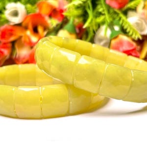 May include: Two light yellow jade bracelets. The bracelets are made of rectangular, polished jade stones. The background features blurred red and orange flowers.