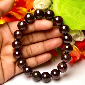 May include: A dark red gemstone bracelet made of round beads. The bracelet is on a hand, with flowers in the background.