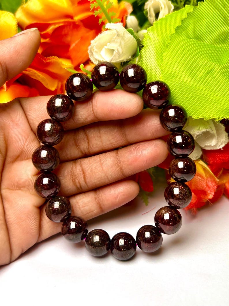 May include: A dark red gemstone bracelet made of round beads. The bracelet is on a hand, with flowers in the background.