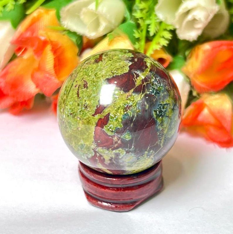 May include: A green and red jasper sphere on a red wooden stand. The sphere is polished and has a smooth surface.