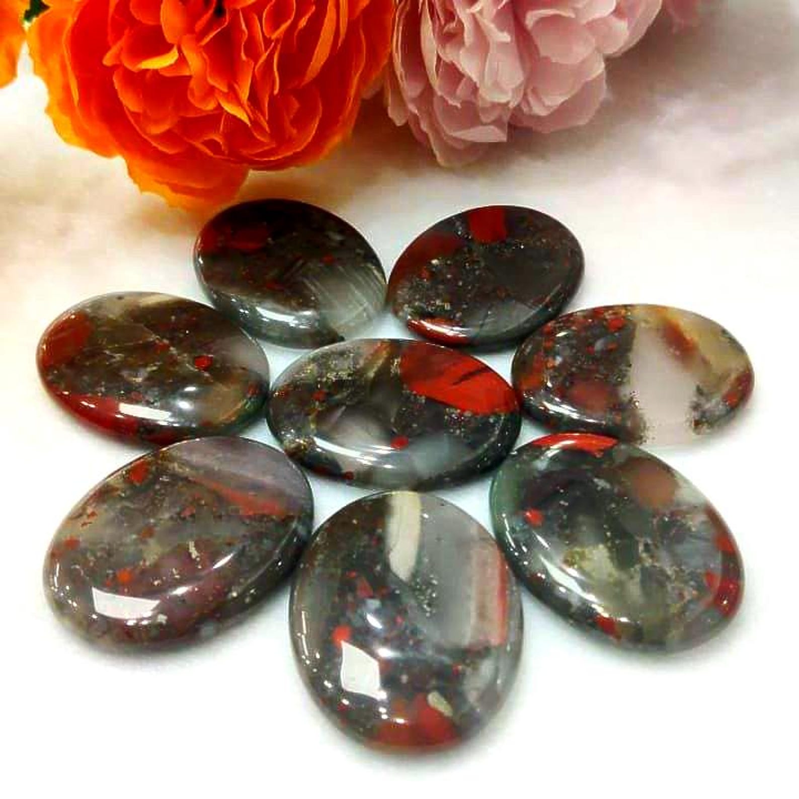Glossy African Bloodstone pocket stones arranged in a circle on white fabric, with orange and pink flowers in background, used for Aries zodiac energy