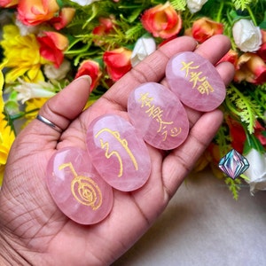 Op de afbeelding: Vier roze rozenkwartsstenen met gouden symbolen erop gegraveerd. De stenen zijn ovaalvormig en glad. De symbolen zijn in Japanse en Chinese karakters.
