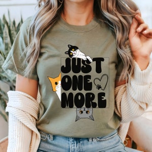 May include: A green t-shirt with a black graphic that says "Just One More" with three cats peeking out from behind the text. The cats are orange, black and white, and gray.