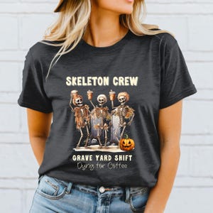 May include: Black t-shirt with a Halloween-themed graphic. The design features three skeleton figures holding coffee cups, with the text "SKELETON CREW" above and "GRAVE YARD SHIFT Dying for Coffee" below. A carved pumpkin is also included.