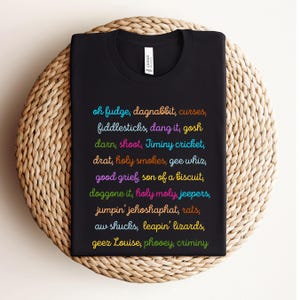 May include: Black t-shirt with colorful text listing various exclamations and phrases. The shirt is folded on a woven, circular mat. The text includes phrases like "oh fudge," "darn," and "good grief."