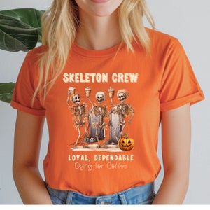 May include: Orange t-shirt with the words "SKELETON CREW" above a graphic of three skeletons holding coffee cups. Below the graphic, the text reads "LOYAL, DEPENDABLE Dying for Coffee". A jack-o'-lantern is also featured.