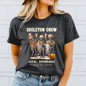 May include: Dark gray t-shirt with a Halloween-themed graphic. The design features three skeleton figures holding coffee cups, with the text "SKELETON CREW" above and "LOYAL, DEPENDABLE Dying for Coffee" below. A jack-o'-lantern is also included.