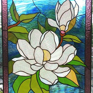May include: A stained glass window depicting two white magnolia flowers with green leaves against a blue background. The window is framed with a pink border.