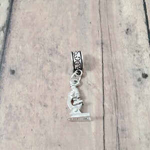 May include: Silver charm featuring a detailed microscope design. The charm is attached to a silver bead with an ornate design.