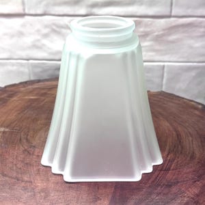 May include: Frosted glass lampshade with a flared, rectangular shape. The shade has a ridged design and a rounded top. The lampshade is sitting on a wooden surface.