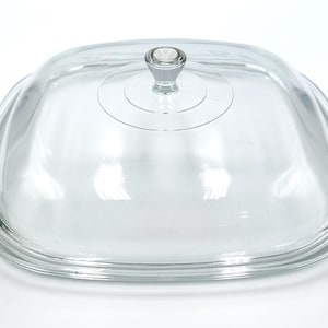 May include: A clear, square glass lid with a small, round knob. The lid has a slightly curved shape and is designed to fit over a square dish. The glass is transparent, allowing visibility of the contents.