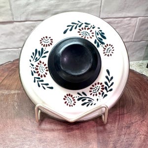 May include: A white ceramic lid with a black knob and floral design in maroon and dark green. The lid is resting on a silver metal stand, set against a wooden surface.
