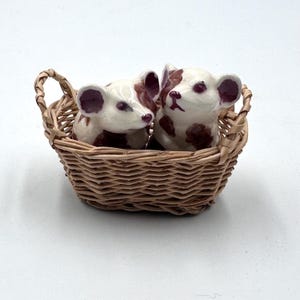 May include: A pair of ceramic mice with brown spots and accents sit inside a small woven basket. The mice have white bodies, brown ears, and dark eyes. The basket is made of light brown wicker.