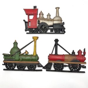 May include: Three decorative train models in red, green, and brown. The trains have black wheels and are set against a white background. The trains are vintage-style and appear to be made of metal.
