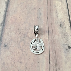 May include: Silver charm featuring a horseshoe with a four-leaf clover in the center. The charm has a decorative band around the top.