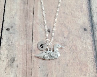 May include: A silver necklace with a loon charm and a small round charm with the letter "C" on it. The necklace is on a wooden background.