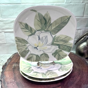 May include: A stack of white ceramic plates with a floral magnolia design. The plates feature a hand-painted magnolia flower with green leaves. The plates are on a wooden surface.