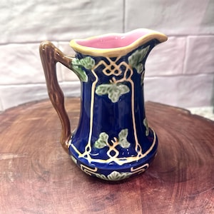 May include: A small, decorative pitcher with a dark blue body, brown handle, and a pink interior. The pitcher features green leaf and vine designs with gold accents. The rim is yellow. The pitcher is likely ceramic.