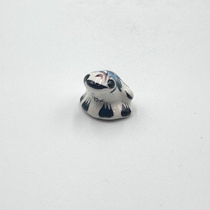 May include: A small, smooth, white ceramic figurine shaped like a frog. The surface features hand-painted dark blue and black abstract markings along the base and sides, with subtle brown and blue accents on the top of the head and back.