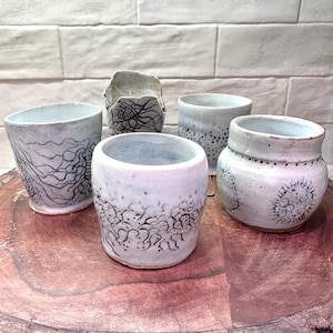 May include: A collection of handmade ceramic cups and a small bowl. The cups are white with unique black line and dot patterns. The bowl is a rough, textured gray. The items are arranged on a wooden surface.