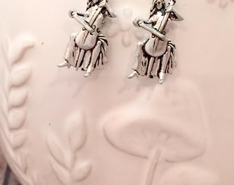 May include: Silver earrings featuring a small figurine of a person playing a cello. The earrings are hanging from a white ceramic bowl with a floral design.