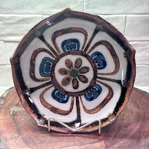 May include: A decorative ceramic bowl with a floral design. The bowl features a white interior with brown, blue, and green floral patterns. The rim is a dark brown color. The bowl is displayed on a metal stand.