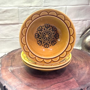 May include: A stack of three ceramic bowls. The top bowl is a warm, golden-yellow color with a scalloped edge and a black floral design in the center. The bowls are stacked on a wooden surface.