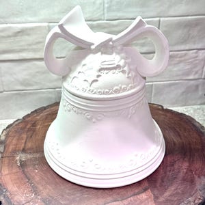 May include: A matte white ceramic bell-shaped container featuring a lid topped with a sculpted bow and holly leaf details. The body of the bell is decorated with embossed scrollwork patterns around the midsection and the bottom rim.