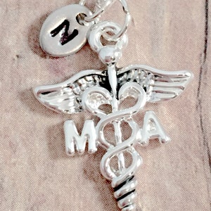 May include: Silver necklace with a medical caduceus charm and a small initial charm. The caduceus charm has the letters "MOA" on it.