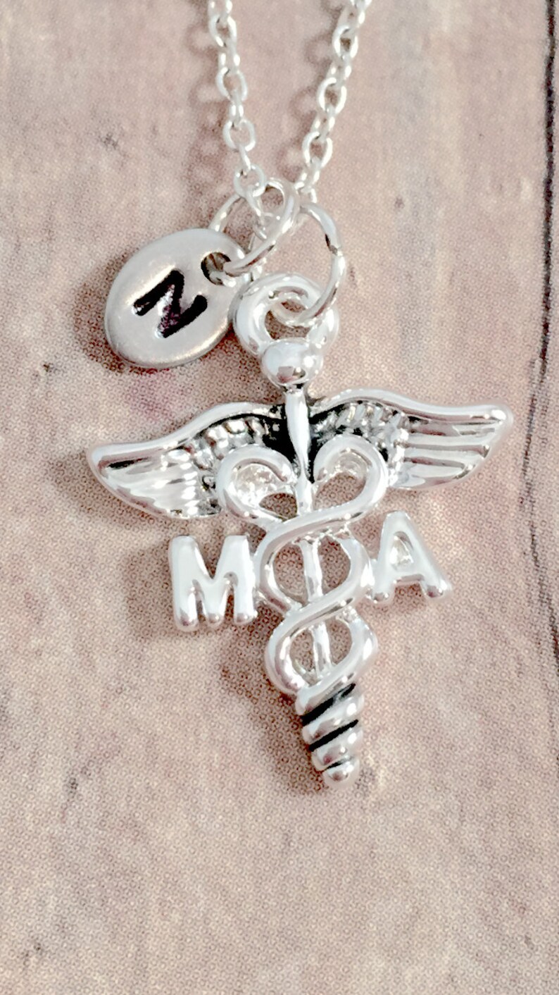 May include: Silver necklace with a medical caduceus charm and a small initial charm. The caduceus charm has the letters "MOA" on it.
