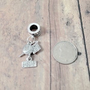 May include: Silver charm with a ball of yarn, crochet hook, and a small sign that says "I love 2 crochet". The charm is attached to a silver bead with a hole for a bracelet.