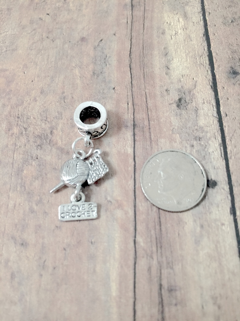 May include: Silver charm with a ball of yarn, crochet hook, and a small sign that says "I love 2 crochet". The charm is attached to a silver bead with a hole for a bracelet.