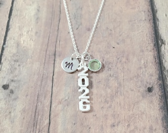 May include: A silver necklace with a charm that says "2026" and a small charm with the letter "M" and a green gemstone.