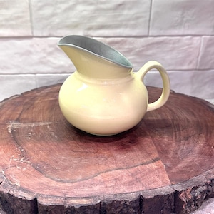 May include: A small, yellow ceramic pitcher with a rounded body and a curved spout. The interior of the spout is a contrasting gray-blue. The pitcher has a single, rounded handle. It sits on a dark brown, circular wooden surface.