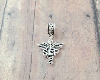 May include: Silver charm with a medical caduceus symbol and the letters "SLP" on a wooden background.