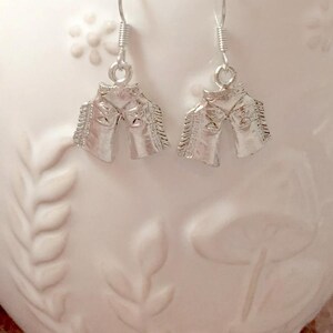 May include: Silver horse head earrings with a detailed design. The earrings are hanging from a white surface.