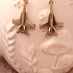 May include: A pair of gold-colored airplane earrings. Each earring features a detailed miniature jet plane charm suspended from a gold-tone ear wire. The earrings are displayed against a white background.