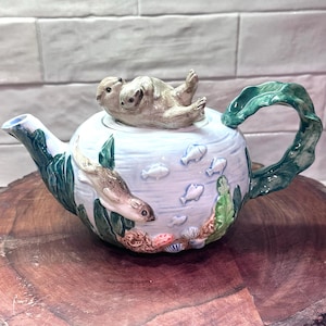 May include: A ceramic teapot featuring a light blue body with painted fish and a sculpted otter swimming along the side. The lid is topped with a sculpted figure of two otters resting together. The handle and spout are shaped like green kelp.