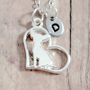 May include: Silver necklace with a heart-shaped pendant featuring a silhouette of a chihuahua. The pendant is attached to a silver chain and has a small silver disc with the letter "D" engraved on it.