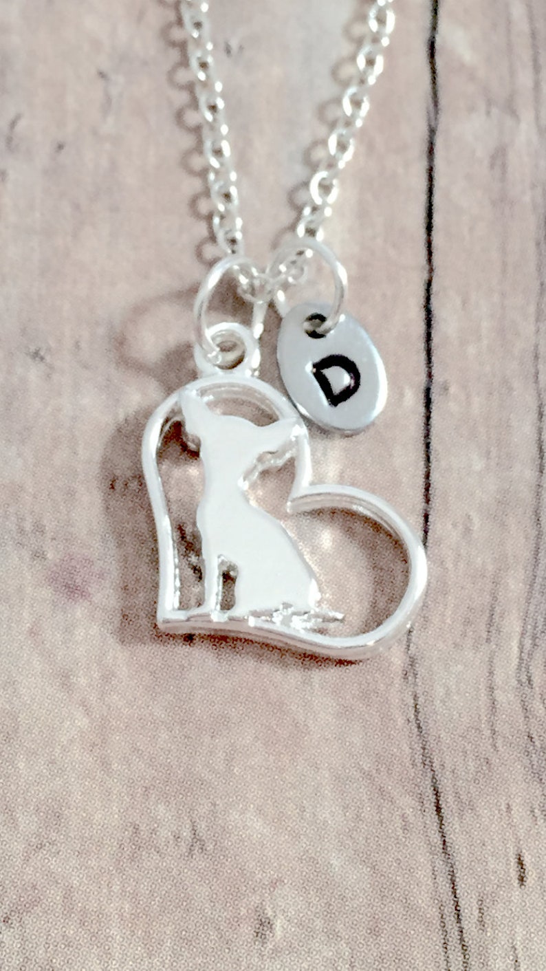 May include: Silver necklace with a heart-shaped pendant featuring a silhouette of a chihuahua. The pendant is attached to a silver chain and has a small silver disc with the letter "D" engraved on it.