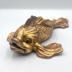 May include: A decorative fish figurine featuring a metallic gold finish with distressed, dark brown accents. The sculpture displays detailed scales, prominent fins, and two inset, glossy, amber-colored eyes, capturing a swimming posture.