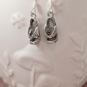 May include: A pair of silver-colored sandal earrings. Each earring features a miniature sandal design with straps and buckles, suspended from a simple hook. The earrings are displayed against a white background.