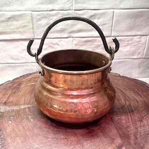 May include: A copper-colored cauldron with a hammered texture and a dark metal handle. The small pot has a rounded body and is sitting on a wooden surface. The handle is arched and attached to the sides of the pot.