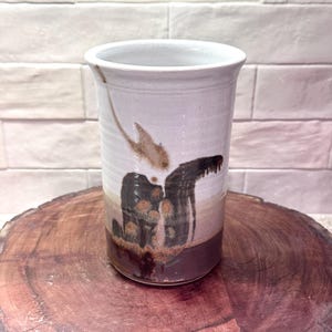 May include: A cylindrical ceramic vase featuring a white upper section with a brown drip glaze pattern and a solid dark brown base. The vessel has a smooth, flared rim and a vertical orientation, showcasing a blend of earthy tones and textures.