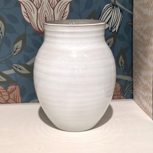 May include: A white ceramic vase with a rounded body and a slightly flared rim. The surface features subtle horizontal ridges throughout the exterior, and the rim has a thin, brownish-red accent line along the top edge of the vessel.