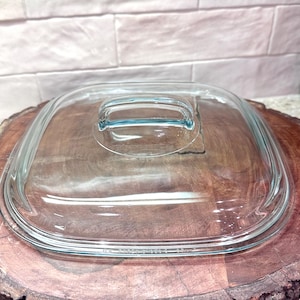 May include: A clear, square glass lid with a rounded handle. The lid is designed for a casserole dish. The glass has a slight blue tint and is set on a wooden surface.