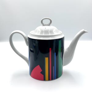 May include: A white ceramic teapot with a black base decorated with colorful geometric shapes and vertical lines. The teapot has a white lid, handle, and spout. The design includes red, yellow, green, blue, and purple accents.