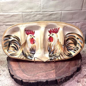 May include: An oval ceramic serving dish with three divided sections, featuring hand-painted illustrations of two roosters with red combs and wattles, brown and black plumage, and cream-colored bodies, accented by faint cursive script text.