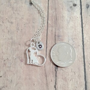 May include: Silver necklace with a heart-shaped pendant featuring a silhouette of a chihuahua. The pendant has a small charm with the letter "D" on it. The necklace is displayed next to a US dime for size comparison.